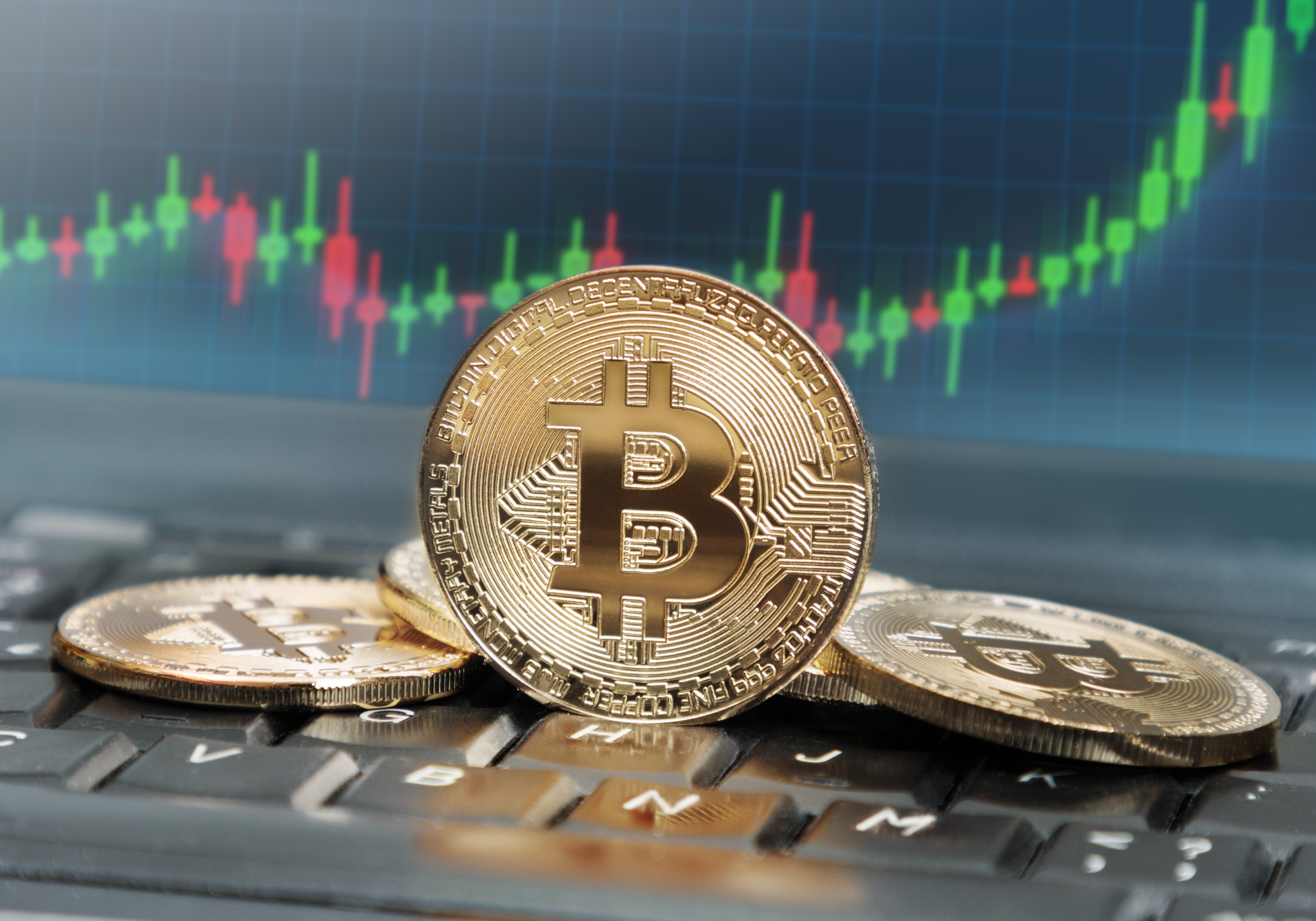 Bitcoins on keyboard with screen in the background displaying rising trend of its value