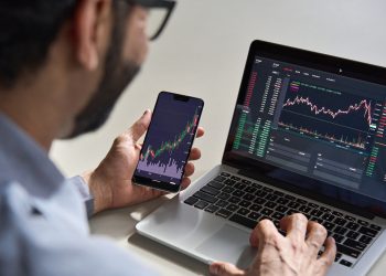 Business man trader investor analyst using mobile phone app analytics for cryptocurrency financial market analysis, trading data index chart graph on smartphone and laptop screen. Over shoulder view